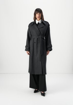 Manteau trench en laine gris avec un devant croisé, taille ceinturée, revers larges et manches retroussées. Associé à un pantalon ample noir.