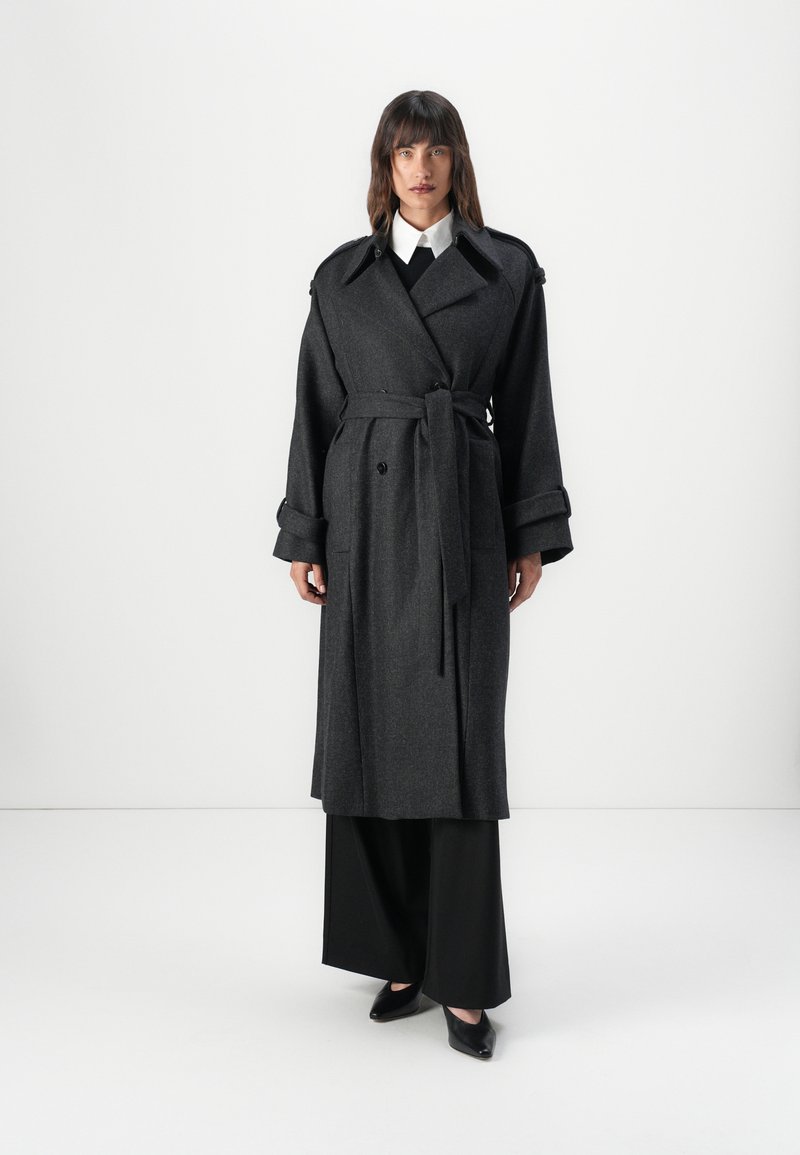 Grau wollener Trenchcoat mit doppelt breiter Front, Taillengürtel, breiten Revers und hochgekrempelten Ärmeln. Kombiniert mit schwarzen weit geschnittenen Hosen.