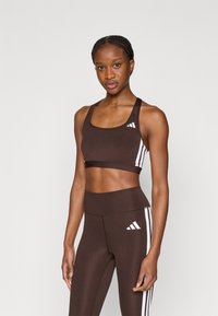 Καφέ αθλητικό σουτιέν με λευκές ρίγες ως διακριτικό, κατασκευασμένο από ελαστικό ύφασμα. Συνοδεύεται από καφέ κολάν με ψηλή μέση που ταιριάζει, και τα δύο με λογότυπα.