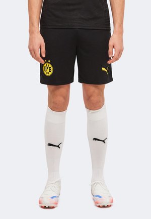 Person trägt schwarze Borussia Dortmund Fußballshorts, weiße Puma Kniestrümpfe und weiße Stollenschuhe mit farbigen Akzenten, steht mit den Händen seitlich.