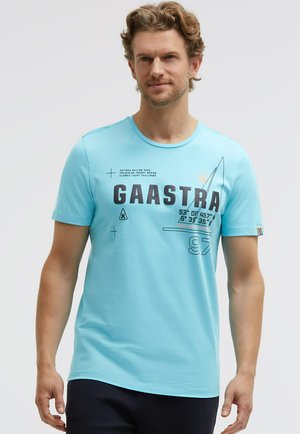 Mann trägt ein hellblaues Gaastra-T-Shirt mit segelthemenbezogenem Text und Grafiken, steht vor einem schlichten hellen Hintergrund.