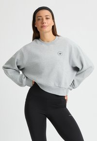 Sudadera gris corta con un cuello redondo y puños de canalé. Detalle del logo negro impreso en el pecho. Combinada con legging negros.