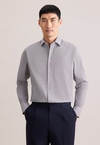 Camicia con bottoni a quadretti piccoli bianchi e blu navy, con colletto, maniche lunghe e taschino sul petto. Indossata con pantaloni blu navy.