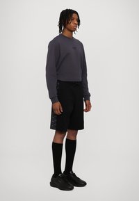 Felpa grigia con logo, pantaloni corti neri con pannello laterale a fantasia, calze nere fino al ginocchio e sneakers nere. Outfit atletico casual.