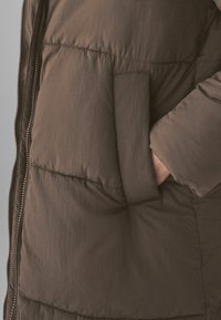Veste matelassée marron avec fermeture éclair, dotée d'une grande poche latérale, texture lisse et détails de couture subtils.