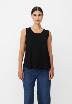 Donna con capelli scuri raccolti in uno chignon elegante, che indossa un top nero senza maniche e jeans blu, in piedi davanti a uno sfondo bianco uniforme.