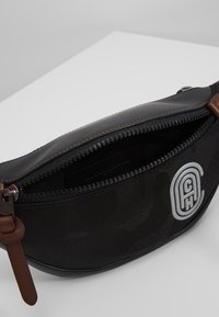 Černý látkový ledvinkový batoh s maskáčovým vzorem, zipovým uzávěrem, hnědými koženými akcenty a výrazným šedým logem na boku.