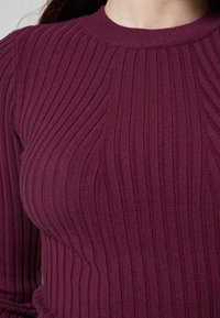 Primo piano di una persona che indossa un maglione a coste, a maniche lunghe, di un viola scuro con un collo rotondo.