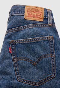 Μπλε τζιν από τζιν ύφασμα με ένα καφέ δερμάτινο ταμπελάκι με τη σήμανση "Levi Strauss & Co." και πορτοκαλί ραφές λεπτομερειών· διαθέτει χαρακτηριστικό σχέδιο πίσω τσέπης.