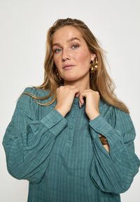 Tyrkysová košile na knoflíky s texturovanými vertikálními pruhy, nabíranými rukávy a mandarínským límcem. Doplňuje ji detail rozděleného manžetu.