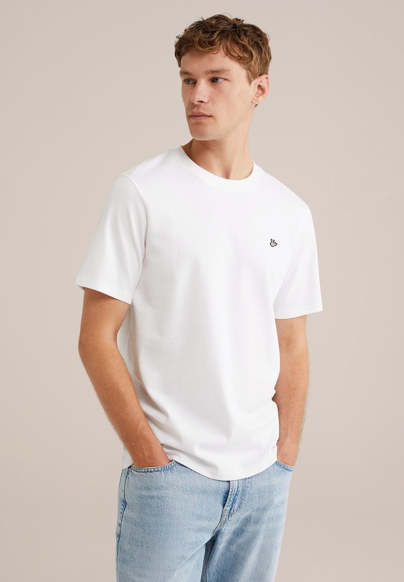 Mlad moški v belem kratkoročnem majici z majhnim logotipom na prsih in svetlo modrih kavbojkah, stoji s rokami v žepih in gleda desno.