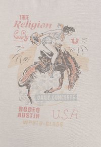 Witte stof met een vintage rodeo-afbeelding van een cowboy op een stampend paard, tekst in rood, blauw en oranje, met een verweerde uitstraling.