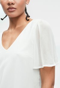 Λευκό πουκάμισο με V-λαιμό και κοντά, διάφανα μανίκια σε στυλ φτερού. Απαλή υφή υφάσματος, με μινιμαλιστικό σχέδιο.