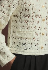 Witte gehaakte cardigan met bloemenpatronen, voorzien van gouden knopen en een zak. De textuur is ingewikkeld, met open brei-details.