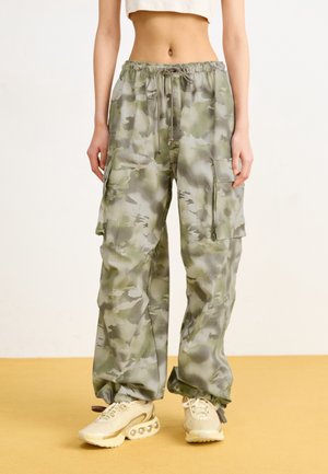 Person wearing loose camouflage cargo pants with drawstring waist and beige sneakers, standing on a yellow floor against a plain background.