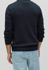 Uomo vestito con un maglione blu scuro e jeans azzurri, visto di spalle su uno sfondo chiaro.