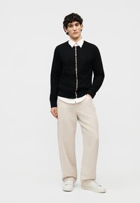 Man wearing black cardigan with multicolor stripe down front, white collared shirt, beige wide-leg pants, and white sneakers, standing with hand in pocket.