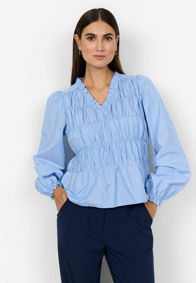 Blusa a righe blu chiaro con scollatura a V, corpetto arricciato e maniche lunghe a palloncino con polsini elasticizzati. Abbinata a pantaloni scuri.