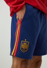 Morski sportski šortsevi s crvenim i žutim bočnim trakama i logom Španjolske nogometne federacije. Mekani pamučni materijal s džepovima.