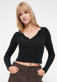 Crop top lung cu maneci lungi, de culoare neagră, cu decolteu în V și închidere cu nasturi. Materialul pare moale, cu un design ajustat. Este băgat în pantaloni maronii.