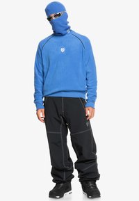 Sweatshirt en polaire bleu avec un col côtelé, pantalon noir ample avec des coutures blanches, bottes noires et un cache-cou en tricot bleu. Lunettes de soleil incluses.
