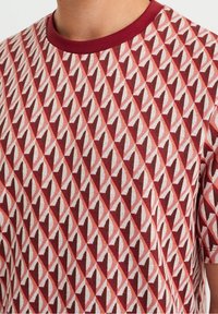 Röd och beige T-shirt med ett geometriskt diamantmönster, med en helt röd rund hals och korta ärmar. Mjuk tygtextur.