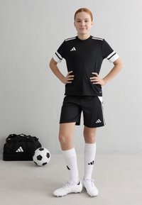 Melnā sporta krekls ar baltām svītrām, atbilstošas šortus, baltas futbola zeķes un baltas futbolbumbas. Melnā sporta soma un futbola bumba tuvumā.