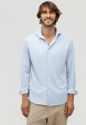 Hombre con cabello castaño medio sonríe, vistiendo una camisa azul clara con botones y mangas remangadas y pantalones beige sobre un fondo liso.