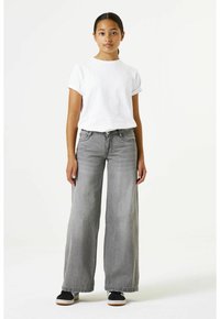 Witte katoenen T-shirt met opgerolde mouwen, gecombineerd met hoog uitgesneden, wijd uitlopende grijze spijkerbroek. Zwarte sneakers met witte zolen.