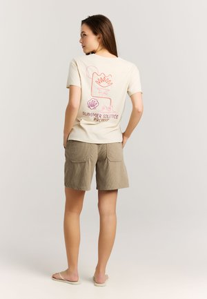 Frau trägt beigefarbene Shorts und ein offwhite T-Shirt mit rotem Linienmuster und dem Text "Summer Solstice Protest" auf der Rückseite, steht mit den Händen in den Taschen.