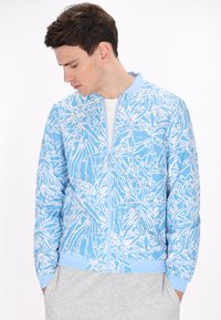 Jachetă bomber bleu deschis cu un model floral abstract alb, manșete și tiv cu nervuri, și închidere cu fermoar frontal. Textură a materialului moale.