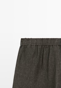 Détail de la ceinture d'un pantalon en tissu élastique marron foncé avec couture froncée sur fond blanc uni.