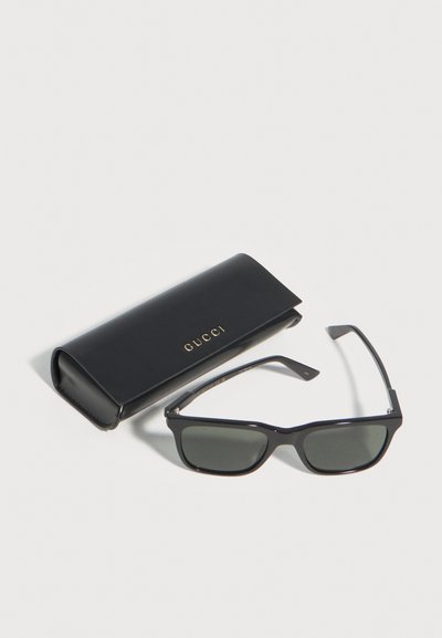 Lunettes de soleil rectangulaires noires avec verres foncés, posées à côté d'un étui Gucci noir avec lettrage doré sur un fond blanc.