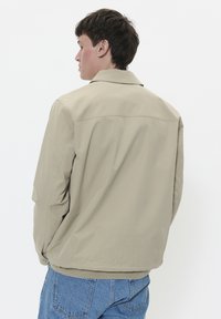 Geacă bej cu o croială lejeră, având guler, cusături pe umeri și talie elastică. Asortată cu blugi din denim albastru.