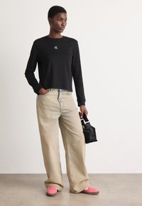 Schwarzes, kurzes Langarm-Shirt mit türkisfarbenem CK-Logo, kombiniert mit hellbeigen, weit geschnittenen Jeans und rosa Sneakers. Accessoire: kleine schwarze Tasche.