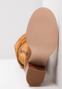 Botín de ante marrón claro con punta redondeada, cremallera lateral y una suela elegante y suave que presenta un tacón con textura de madera y fondo acanalado para mayor agarre.