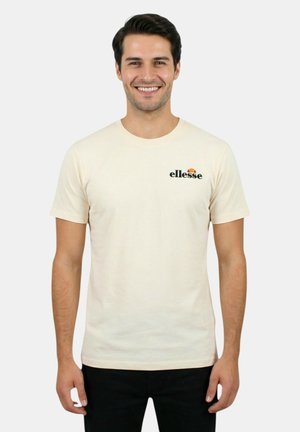 Uomo sorridente che indossa una t-shirt Ellesse color crema e pantaloni neri, in piedi davanti a uno sfondo grigio chiaro uniforme.