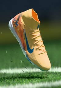 Chuteira de futebol Nike amarela e laranja com logo azul, no ar acima de um campo de relva, com os cravos da chuteira e partículas de relva visíveis.