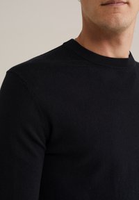 Primer plano de una persona que lleva un suéter negro de cuello redondo liso, mostrando el cuello, el hombro y parte de la parte inferior de la cara.