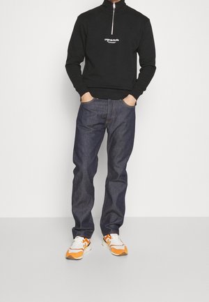 Person trägt einen schwarzen Sweatshirt mit Viertel-Reißverschluss und dem Schriftzug "Originals Studio", blaue Jeans und weiße Sneaker mit orangefarbenen Akzenten.