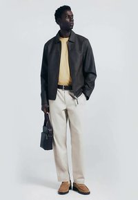 Mörkbrun läderjacka över en gul skjorta, kombinerad med cremefärgade byxor och bruna skor. Bär en svart väska. Enkel, strukturerad design.