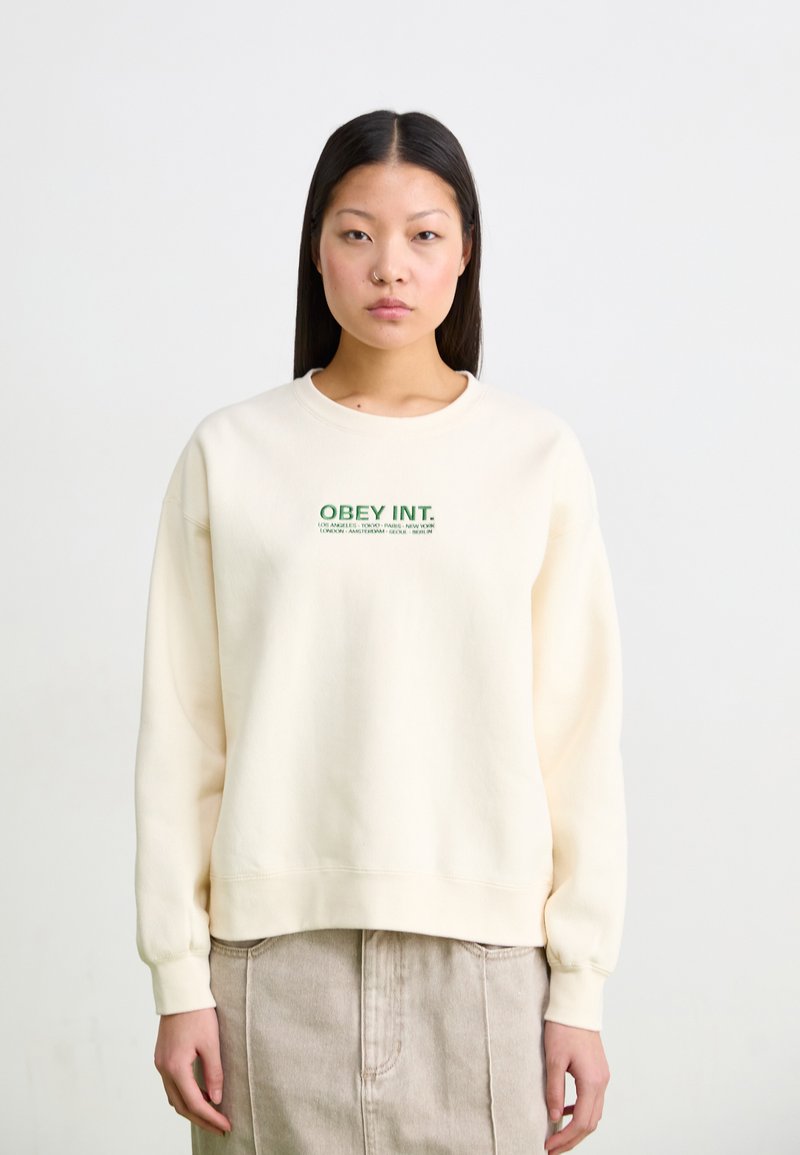 Cream-colored sweatshirt with a round neck and ribbed cuffs, featuring green "OBEY INT." text across the chest. Soft fabric texture.