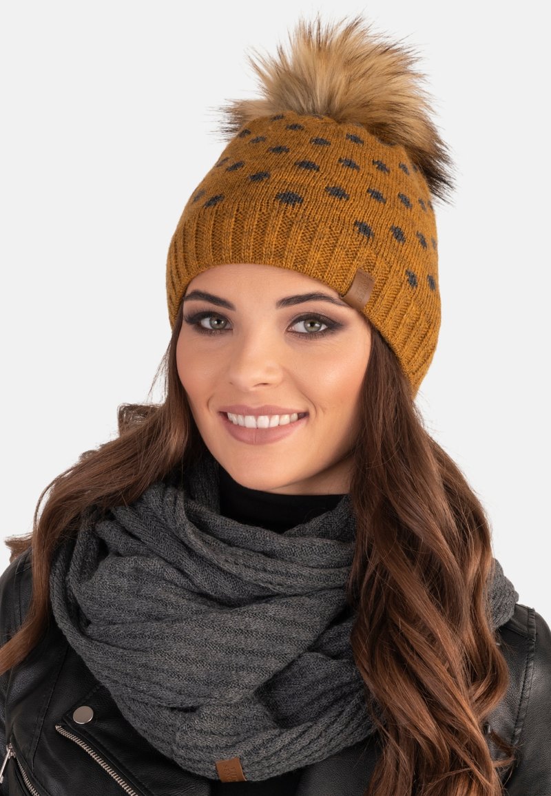 Knitted mustard beanie with black polka dots and a fur pom-pom on top. Paired with a gray ribbed scarf.