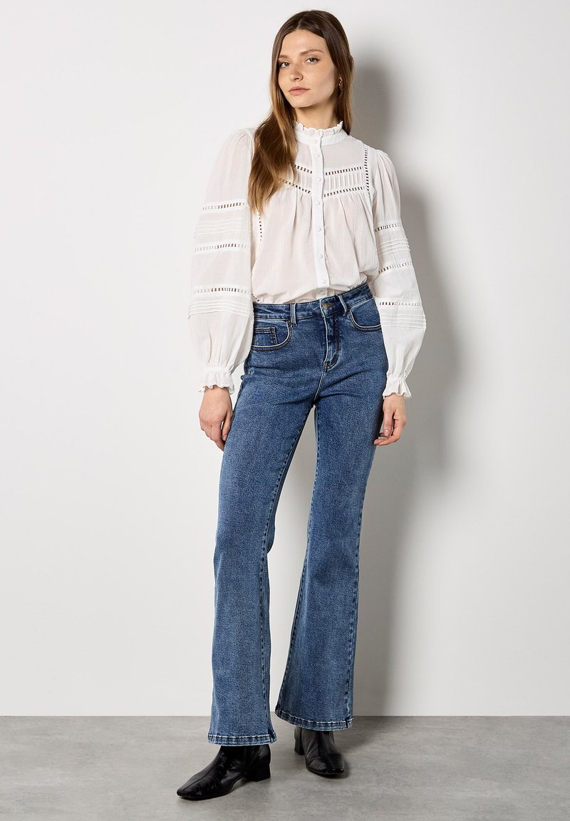 Jeune femme portant un chemisier blanc à manches longues avec des détails en dentelle et un jean bleu évasé taille haute, debout devant un fond uni.