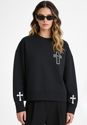 Frau trägt schwarzen Pullover mit weißen Kreuzmotiven an Ärmeln und Brust sowie schwarze Sonnenbrille, steht vor weißem Hintergrund.
