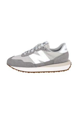 Zapatilla New Balance gris y blanca con superposiciones de ante, logo "N" blanco, cordones grises y suela de goma marrón, mostrada de lado.