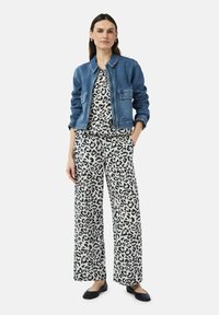 Veste en jean, haut imprimé léopard et pantalon large blanc avec un motif noir. Modèle portant des ballerines noires, les mains dans les poches.