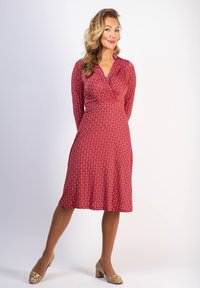 Punainen, kuvioitu mekko V-pääntiellä, pitkät hihat ja liehuva helma. Yhdistetty beigen värisiin korkokenkiin, joissa on kultaisia yksityiskohtia. Sileä kankaan pinta.