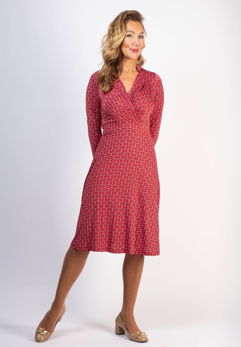 Punainen, kuvioitu mekko V-pääntiellä, pitkät hihat ja liehuva helma. Yhdistetty beigen värisiin korkokenkiin, joissa on kultaisia yksityiskohtia. Sileä kankaan pinta.