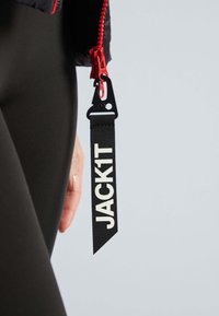 Schwarzes Stoffetikett mit "JACK1T" in Weiß, befestigt an einem roten Reißverschluss einer Jacke. Glatte Textur, geradliniges Design, mit einer Schlaufe zum Befestigen.